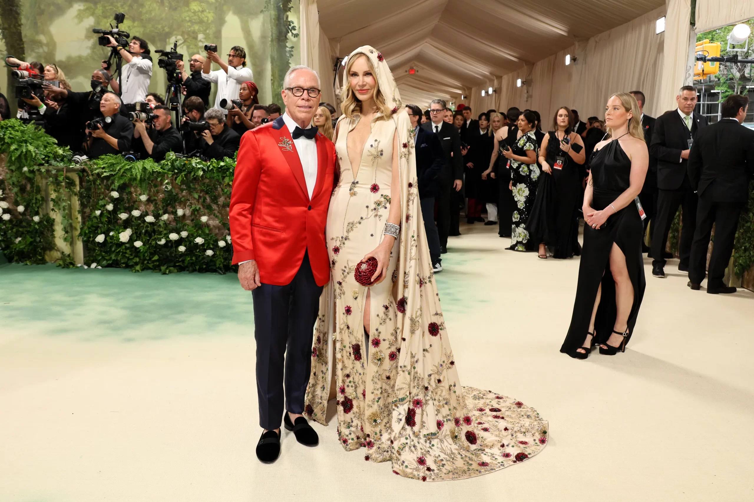 Tommy Hilfiger in een pak met rode blazer en Dee Ocleppo in een nude jurk met rode bloemen