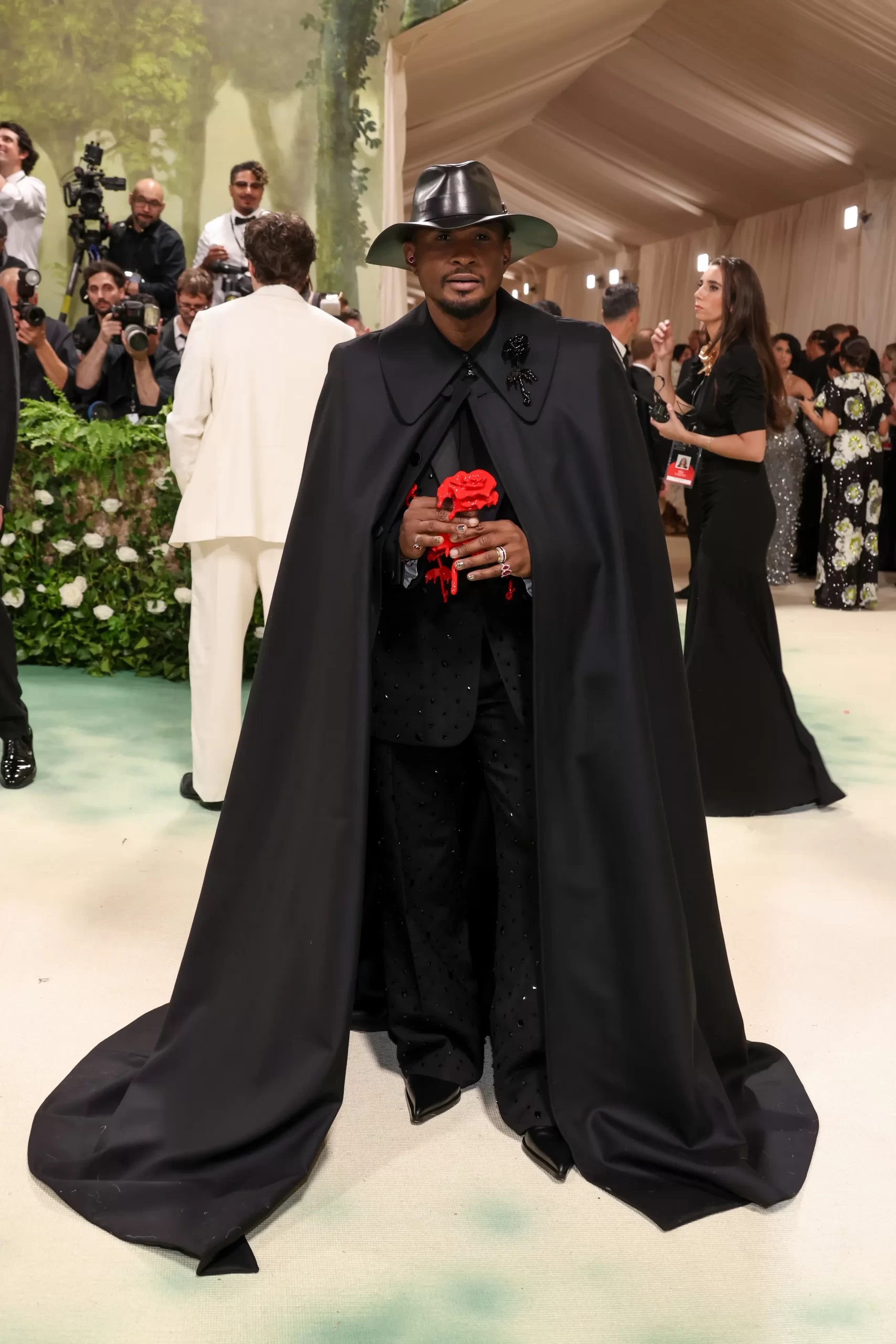 Usher draagt een zwarte look met lange, dramatische cape en een rode roos.