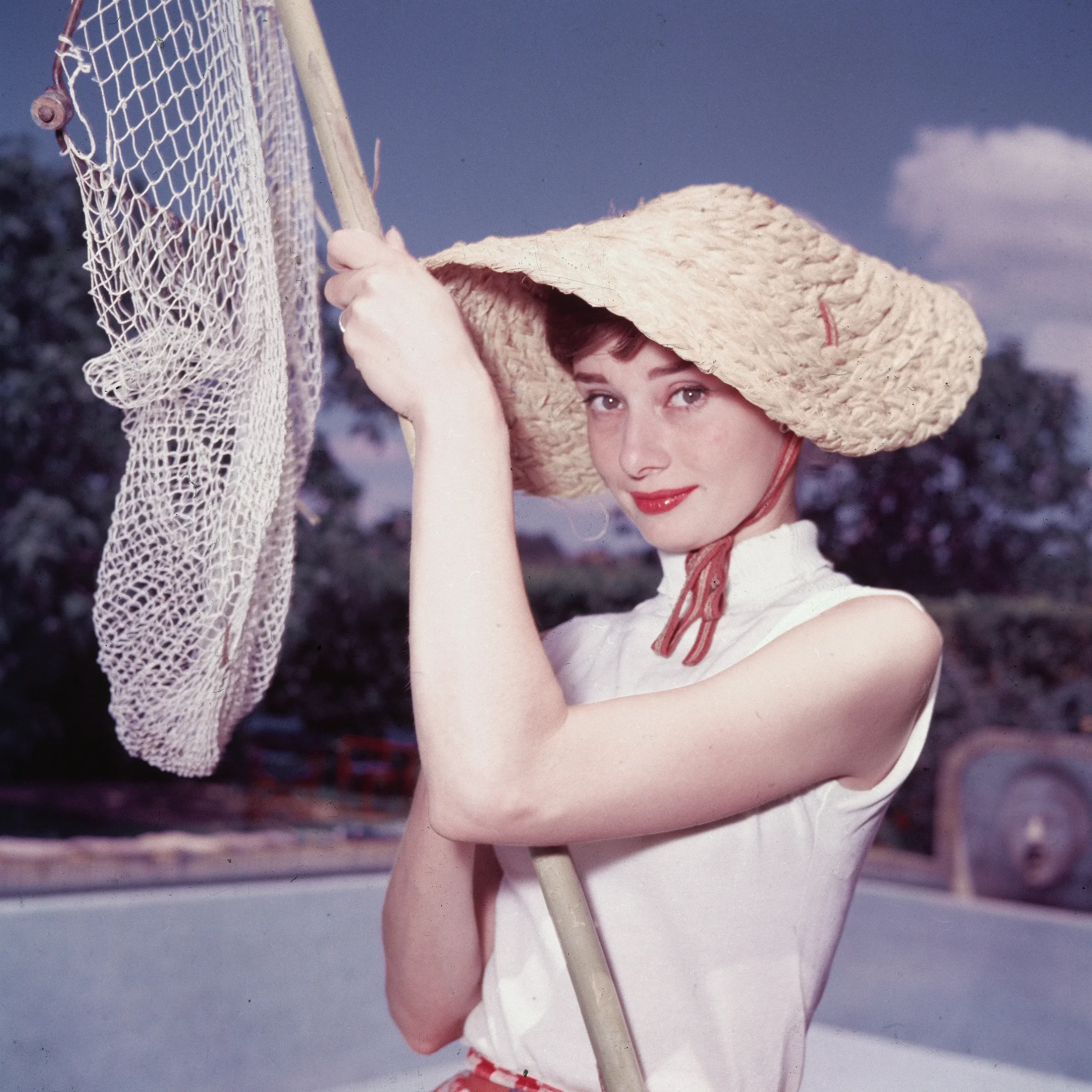 Audrey Hepburn met een zonnehoed en een mouwloze blouse naast een zwembad, begin jaren vijftig.